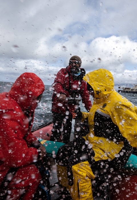 People in bright orange and yellow rain gear in an orange skiff boat through choppy waters through a rain spattered lens. A blue and white ship is in the background.