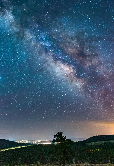 A bright light obstructs the night sky and milkyway directly over a city at night with hills and trees in the foreground and stars pictured in the background.