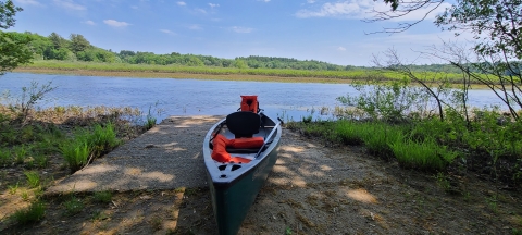 Photo of the canoe launch off of Weir Hill Trail, with a dark blue canoe sitting on the ground in front of the Sudbury River