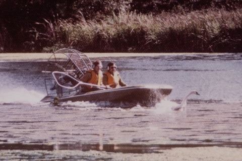 an old photo with faded color shows two men in a boat wearing life jackets. One of the men is holding a net. They boat is inches from a fleeing swan.