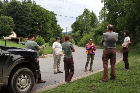 A group of people stand at a road's edge, one person speaks to the rest of the group
