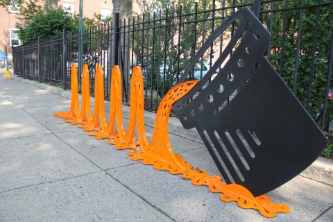 a metal art installation of orange waves pouring from a black can sits on a sidewalk, with a black metal fence and green shrubs in the background