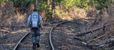 A person wearing a technical backpack walks down railroad tracks in the forest