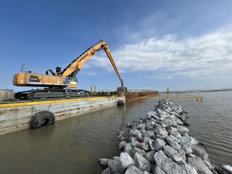 A large mechanical crane on a barge places gray rocks in a line.