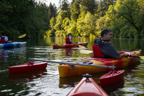Several people kayak on a calm, tree-lined river in warm evening light. In the foreground, a man wearing a red life jacket and a dark plaid shirt sits in a bright yellow and red kayak with red outriggers on either side for stability, while two other kayakers paddle nearby. Dense green trees reflect on the water’s surface.