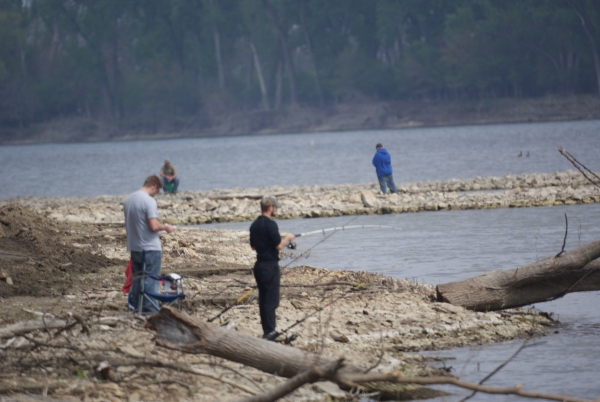 Four fishers along the edge of the water on a rocky bank with dead logs around.