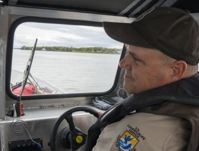 A Federal Wildlife Officer sits at the helm of a boat with the banks of the river visible through the window in front of him.