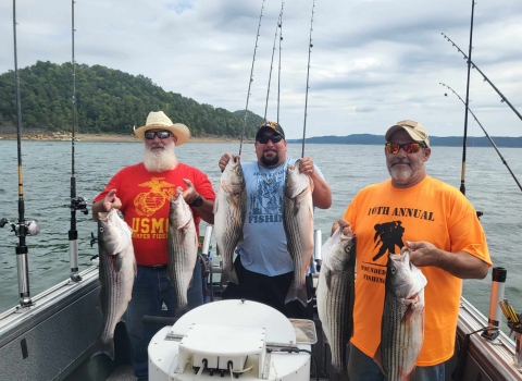 Three men smiling holding a striper in each hand on a boat on a lake