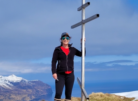 A woman wearing a hat and sunglasses on a high summit overlooking snowcapped mountains and ocean. She stands next to a metal structure.