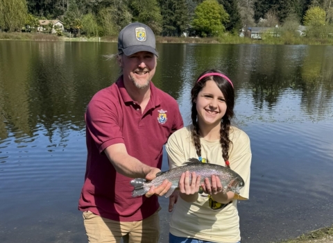 Service staff member and youth event participant posing with a rainbow trout. Lake, houses and trees in the background.