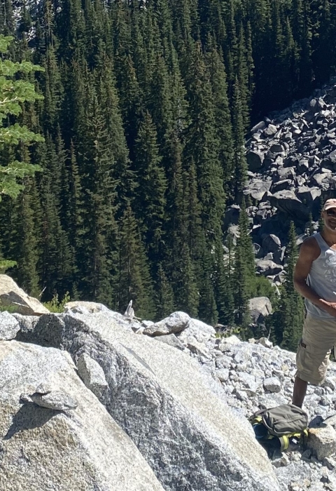 A man stands on the side of a rocky mountain with rocks, trees and greenery around him.