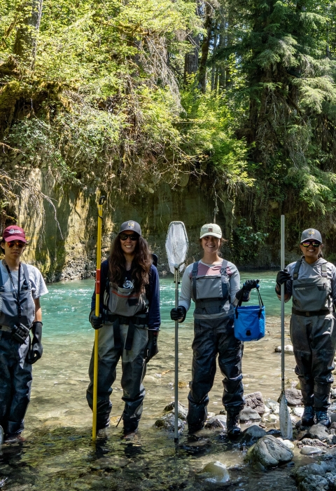 Four people stand at the shore of a river wearing waders and holding fishing nets as they smile at the camera. The shore is rocky, and trees can bee seen on the other side of the river.