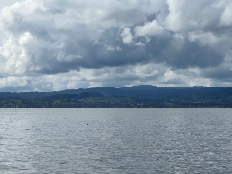 A slightly rippling lake is surrounded by blue and green hills. The sky has large clouds
