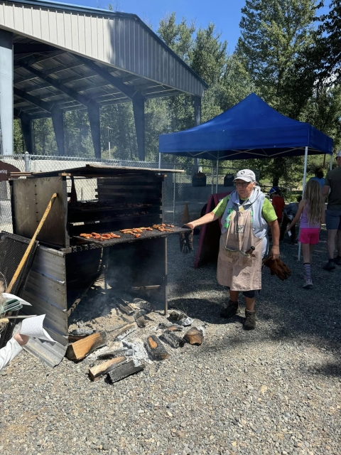 A women pulls out a rack of salmon that is smoking over burnt logs