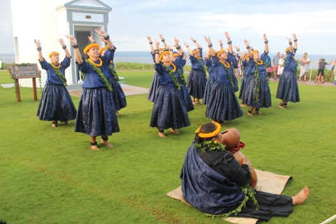 Hawaiian cultural practicioners dance and chant during a mele on a green field by light house at Kilauea Point NWR
