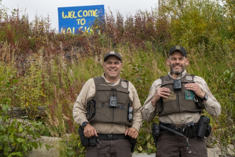 Two uniformed Federal Wildlife Officers pose for a picture with a painted sign in the background on a hill reading "welcome to Kalskag" 