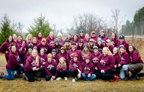 Group of women of varying ages pose for a photo along a roadside while wearing matching maroon sweatshirts made for the trapping workshop. 