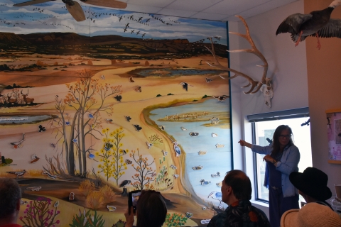 A person gestures towards a wall-covered mural while standing in front of an audience.