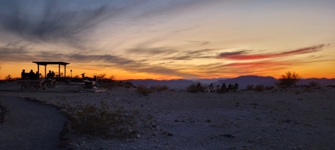 View of sunset on Child's Mountain. Visitors silhouetted sitting on camp chairs, at picnic tables, and around grill in front of ramada. 