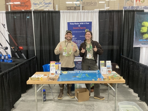 Two biologist stand smiling behind an outreach booth focused on invasive species prevention and identification in Alaska.