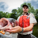 A person in a Service uniform and life vest holds a large hideous fish with bulging eyes and a gaping mouth up the camera. 