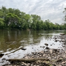 View looking downstream a flowing river from a rocky bank, with trees on the far side.