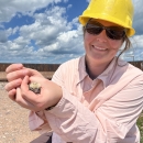 A biologist in a yellow hard hat holding a small fuzzy bird while kneeling on sandy ground