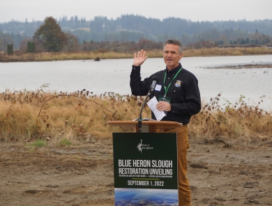 A man in a US Fish & Wildlife Service shirt stands behind a poduum that says "Blue Heron Slough Restoration Unveiling" in front of a body of water