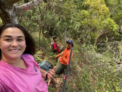 A woman on a steep wooded slope poses for a selfie. Two people smile in the background.