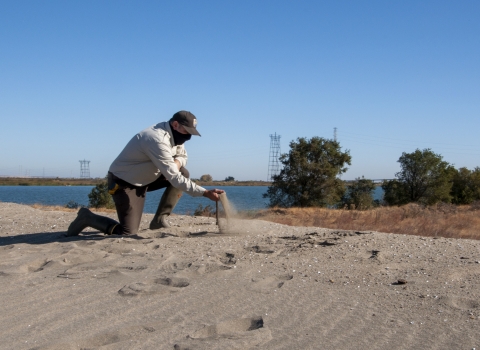 Kneeling man picks up a handful of sand with a body of water in the background