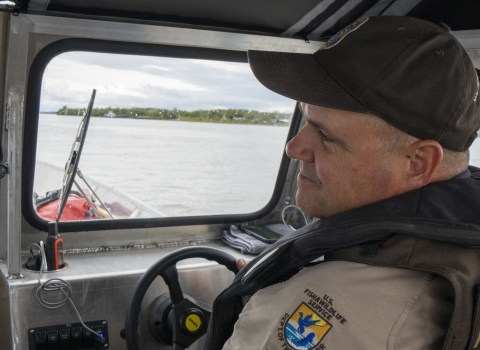 A Federal Wildlife Officer sits at the helm of a boat with the banks of the river visible through the window in front of him. 