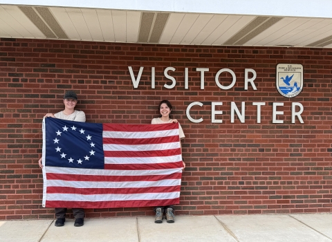 2 people hold up Betsy Ross flag in front of brick wall saying Visitor Center