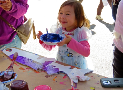 Smiling little girl holds a bowl of blue paint with a partially painted wooden salmon outline on the table before her.