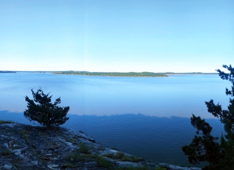 Panoramic view from lichen-covered bluff overlooking blue water and sky with green summer foliage in the distance