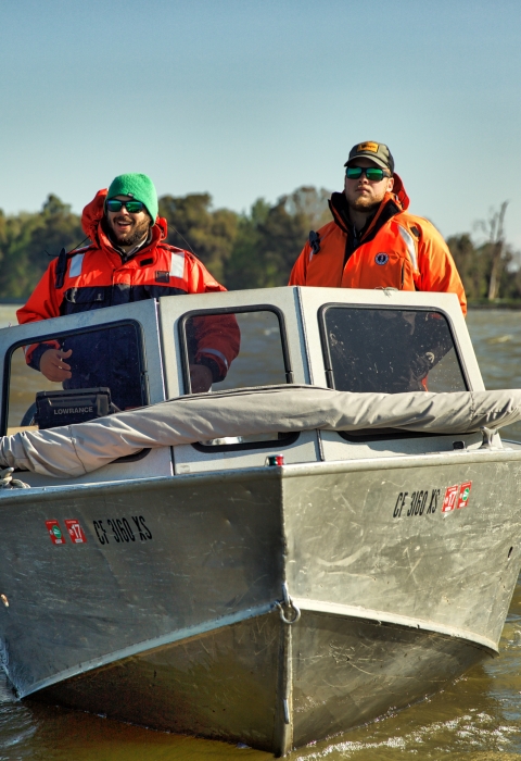 Two men pilot a motored boat on a river