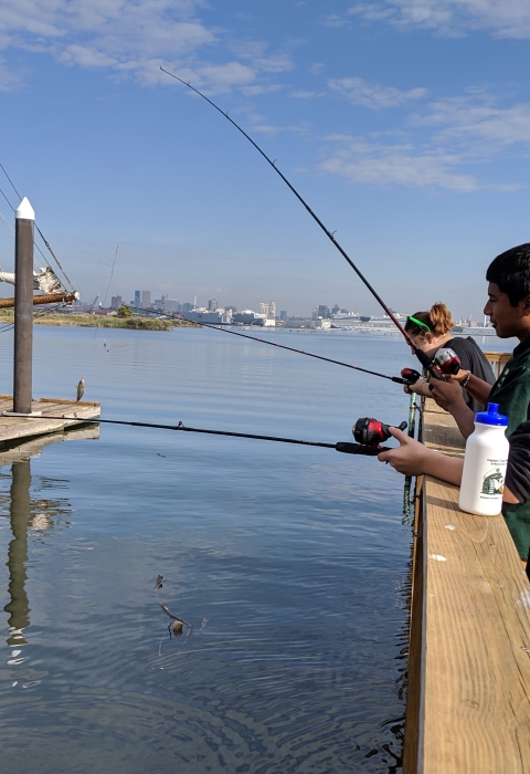 Three students fish from pier at Masonville Cove with Baltimore skyline in background