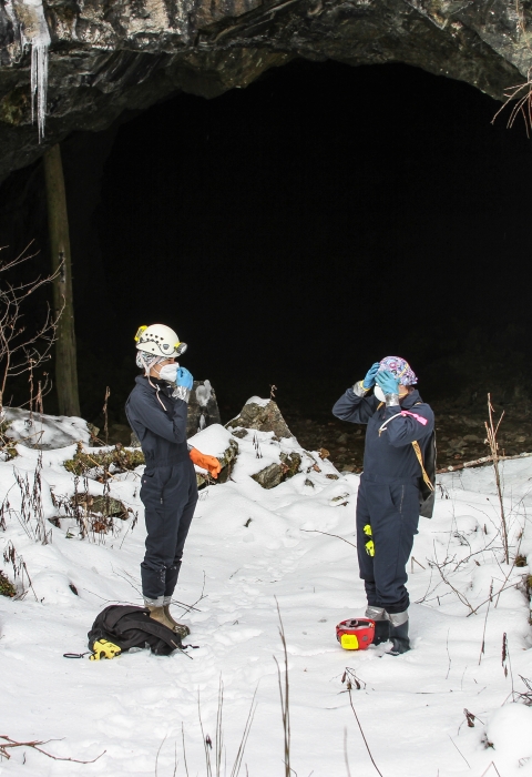 Two people stand in the snow outside a mine entrance, making adjustments to their helmet and face mask