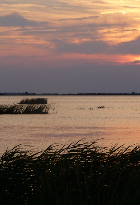 A setting sun ducks behind clouds over an expanse of open water dotted with marsh grasses