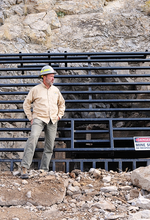 A man in a yellow shirt with a hard hat on stands by a grate covering an opening in the rocks. He is looking over his left shoulder.