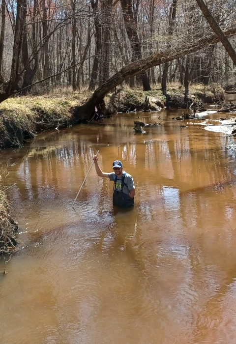 Man in waders walks in murky water surveying water depth