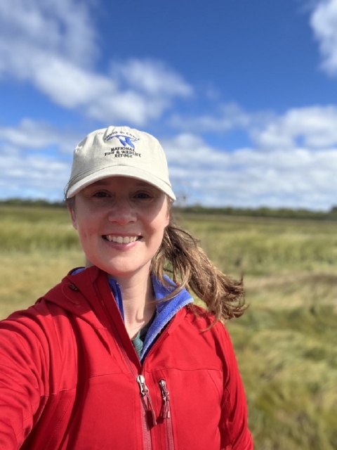 Woman stands in a marsh wearing fish and wildlife hat