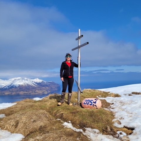 A woman wearing a hat and sunglasses on a high summit overlooking snowcapped mountains and ocean. She stands next to a metal structure and a buoy that sits on the ground.