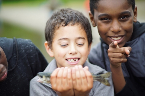A young child looks at a fish he is holding. A child next to him smiles and points at the camera. 