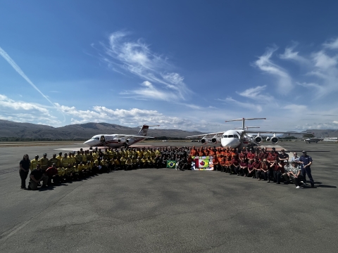 A group of people stand together in a semi circle on a tarmac in front of aircraft used for firefighting efforts. The group has flags out and are all in firefighter clothing or agency t-shirts.