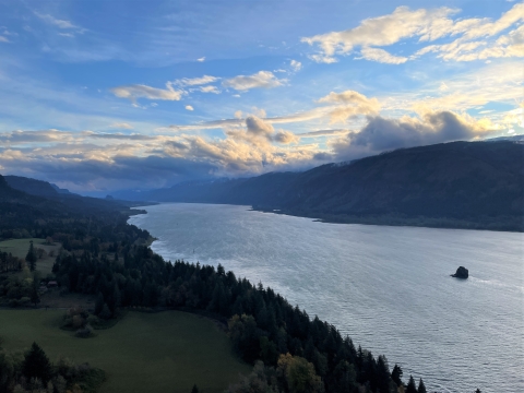 A view of a river from a high point, looking down at the river between hills and mountains with trees. The sky is blue with clouds and low, golden light.