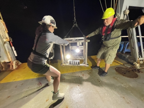 A man and a woman are holding onto a metal structure on the aft deck of the ship. They are bringing the video camera system back up onto the boat. They are both wearing personal floatation devices and hard hats. It is night time.