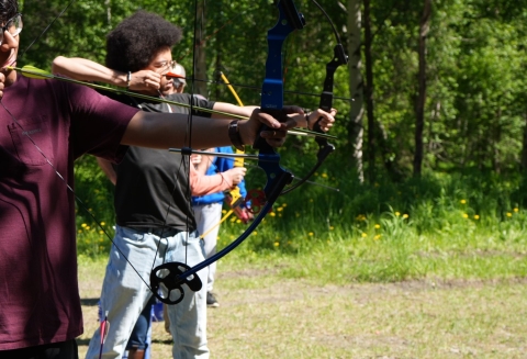 looking down the firing line of an archery range with two kids at full draw with their bows as they aim at the target off the image view. Woodline vegetation in the background. 