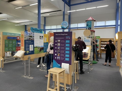 Six people engaging with panels of the We Are Water exhibit at North Branch Area Library. Notable titles of the panels include "Stories from Chisago County" with attached headphones so visitors can listen to people share their stories, "What's in the water?", a water tower that says "Minnesota's drinking water", and "you+me+water".