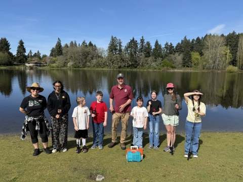 Three adults and six youth with fishing rods and tackle standing on grass in front of a lake. Lake, trees, and sky in the background.