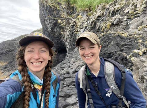 Two biologist smiling while taking a selfie on St Lazaria. Volcanic rock in the background.